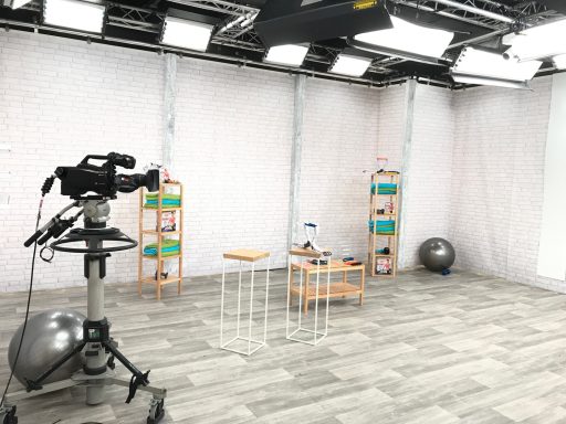 Studio vidéo avec un caméraman, un meuble, et des étagères colorées. Mur blanc.